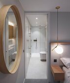 Schlafzimmer mit rundem Wandspiegel neben offener Tür und Blick ins beleuchtete, minimalistische Bad