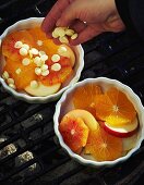 A hand putting white chocolate chips over slices of fruit in small ovenproof gratin dishes