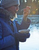 A woman dressed in warm clothing eating soup by the water