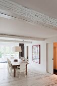 Wooden dining table and antique white upholstered chairs below wood-beamed ceiling