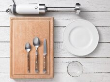Assorted kitchen utensils: a wooden chopping board, cutlery, crockery and a stick blender