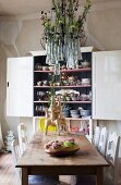 Aufgehängte Glasvasen mit Blumendekoration über Holztisch mit künstlerischen Vasen und vor vollem Geschirrschrank