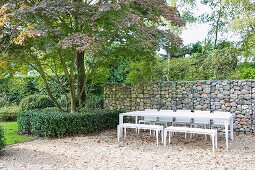 White outdoor furniture on gravel terrace with gabion wall in garden
