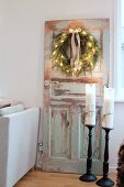 Candles in black candlesticks on floor in front of Christmas wreath with fairy lights on vintage door