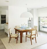 Simple dining set under white lampshade against partition wall with fitted kitchen in background