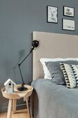 Black lamp on bedside table next to bed with beige headboard and grey cover against grey wall
