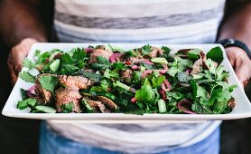 A man is holding a plate of Thai Grilled Beef and Herb Salad