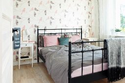 Black metal bed with grey and dusky pink bed linen against wall with bird-patterned wallpaper