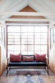 Couch with scatter cushions in bright gable window with chipboard cladding and recycled board floor