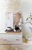 Feminine accessories on tray in front of artworks leaning against wall on top of pale wooden cabinet