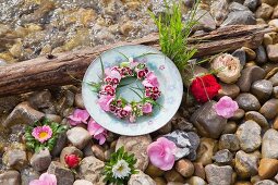 Teller mit Blumenkranz und Blüten im Kiesbett am Wasser