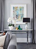 White orchid, vases and Fornasetti table lamp on console table in front of modern picture on gray wall