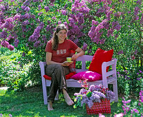 Woman with coffee cup on pink wooden bench in front of flowering Syringa vulgaris