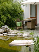 Blick vom Schwimmteich mit Holzsteg auf Terrasse mit Korbmöbeln