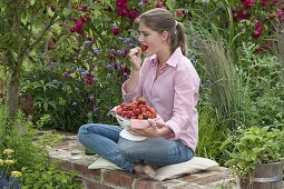Junge Frau sitzt auf Mauer und nascht frisch gepflückte Erdbeeren