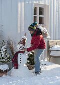 Frau baut Schneemann mit Tontopf als Hut, Schal, Kohlstrunk als Pfeife