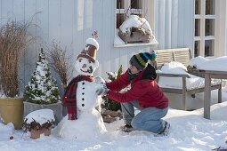 Frau baut Schneemann mit Tontopf als Hut, Schal, Kohlstrunk als Pfeife