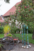 Gravel patio, old cart with wooden wheels, grass and Bergenia (bergenia) in terracotta pots, decorative plugs of handmade pottery, candlesticks, baubles, apple tree (Malus)