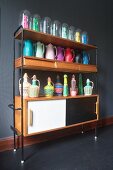 Retro Sideboard mit Regal und verschiedenen bunten Thermoskannen, Korbflaschen und Frauenfiguren unter Glasglocken