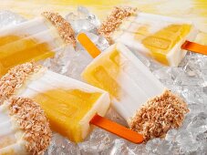 Mango and coconut ice lollies topped with white chocolate and desiccated coconut on a bed of ice cubes (close-up)