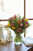 Luxuriant bouquet of tulips and delicate touch-me-not in glass vase next to china rabbits