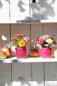 Posies of zinnias in glasses of water wrapped in felt ribbons and apples on white shelf