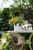 Sunflowers and ornamental grasses in jug on garden table with wicker armchair in background