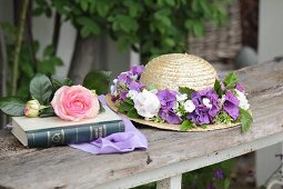 Strohhut mit Blumenkranz neben einem Buch mit Rose im Garten