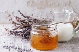 Open glass jar of liquid honey with honeycomb and silver spoon inside, glass jug of milk and bunch of dry lavender
