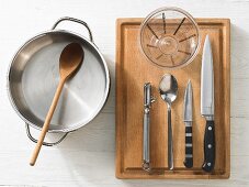 Various kitchen utensils: pot, wooden spoon, measuring cup, peeler, knives