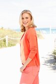 A blonde woman on the beach wearing a T-shirt, cardigan and trousers in different orange tones