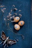 Easter eggs decorated with animal stickers, dried twigs and metal insect figurine on dark blue fabric