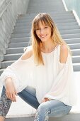 A blonde woman wearing a white blouse and jeans with embroidered flowers, sitting on a flight of stairs