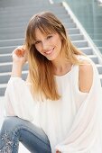 Blonde Frau in weißer Bluse und Jeans mit Blumenstickerei sitzt auf einer Treppe