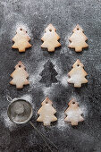 Rows of Christmas tree shaped biscuits dusted with icing sugar with one missing with just the outline remaining and a mini sifter filled with icing sugar all on a grey slate surface