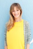 A blonde woman wearing a yellow top and a grey cardigan in front of a blue wall