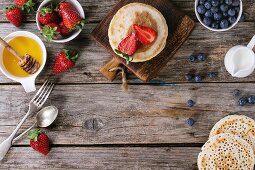 Pancakes with strawberries and blueberries, bowls of honey and jug of milk over old wooden background