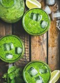 Fresh green smoothie in bottle and glasses with ice cubes, mint and lime in wooden tray, top view