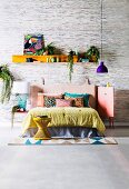 Bunte Decke und Kissen auf Bett mit Lederkopfteil, Pendelleuchte mit lila Lampenschirm und Regal mit Bild und Zimmerpflanzen vor Tapete