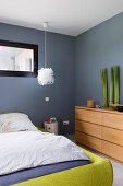 Double bed with green frame and houseplants on chest of drawers in bedroom with grey walls