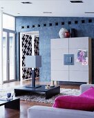 Designer cupboard with felt-covered doors against blue-grey wall, low table on flokati rug, table lamps and hot-pink cushions