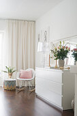 weiße Kommode mit Blumenstrauß und transparenter Tischleuchte, Klassiker-Schaukelstuhl und Beistelltisch vor Fenster mit Vorhang