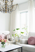 weiße Polstergarnitur und Beistelltisch mit Zimmerpflanze vor Fenster, im Vordergrund Couchtisch mit Rosenstrauß