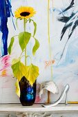 Sunflower in blue vase next to elegant sequinned hat and feminine shoe horn in front of painting