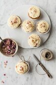 Rose and lemon cupcakes with rosewater and lemon frosting tied with string with spoon and bowl of rose petals, strong and knife on a white and grey marble surface