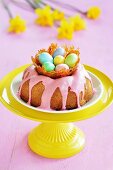 A gugelhupf with a pink sugar glaze and a caramel nest with colourful sugar eggs on a stand, with daffodils in the background