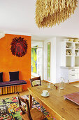 Holztisch mit Stühlen auf buntem Teppich und lila Sitzbank vor orangefarbener Wand
