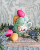 Colourful speckled eggs in gold-rimmed teacup and Eryngium flower on rustic wooden surface