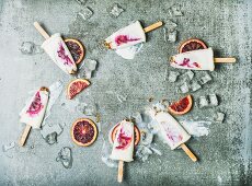 Blood orange, yogurt and granola popsicles on ice cubes over grey concrete background