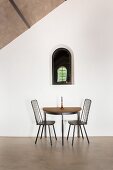 Semicircular table and two chairs below small arched window